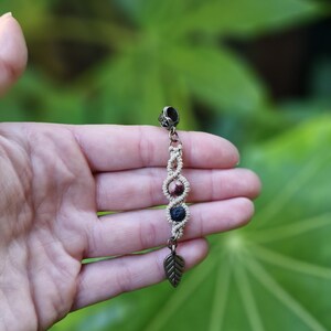Puede incluir: Pendiente de macram&eacute; hecho a mano con un dije de hoja color bronce y cuentas decorativas en tonos marr&oacute;n y azul. El pendiente se muestra sobre un fondo de follaje verde.