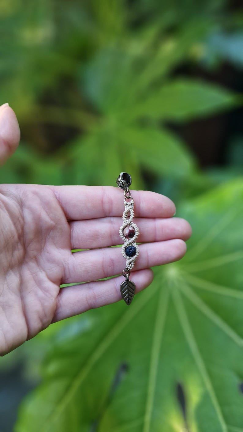 Puede incluir: Pendiente de macram&eacute; hecho a mano con un dije de hoja color bronce y cuentas decorativas en tonos marr&oacute;n y azul. El pendiente se muestra sobre un fondo de follaje verde.