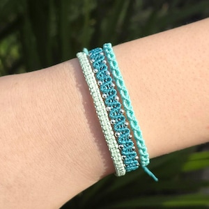 Op de afbeelding: Drie macramé armbanden in tinten van turquoise en mintgroen worden getoond. De middelste armband heeft zilveren kralen, de andere hebben geweven patronen. De armbanden worden om een pols gedragen en tonen de handgemaakte sieraden.