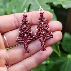 Könnte beinhalten: Ein Paar bordeauxfarbene Makramee-Weihnachtsbaum-Ohrringe mit silbernen Haken. Die Ohrringe sind mit kleinen goldenen Perlen verziert, was einen festlichen und handgefertigten Look erzeugt. Die Ohrringe werden in einer Hand vor einem verschwommenen grünen Hintergrund gehalten.