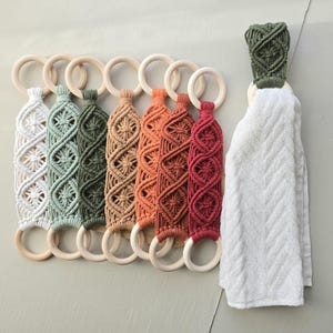 May include: A collection of macrame towel holders in various colors, including white, green, brown, orange, and red. Each holder features a wooden ring and intricate woven patterns. A white towel hangs from one of the holders.