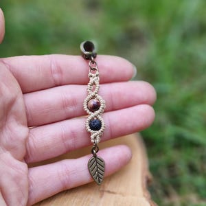 Puede incluir: Un pendiente de macram&eacute; hecho a mano con tres cuentas de piedra en tonos marr&oacute;n y negro. El pendiente presenta un dije de hoja y una parte superior de metal color bronce. El fondo est&aacute; borroso, con follaje verde y una superficie de madera.