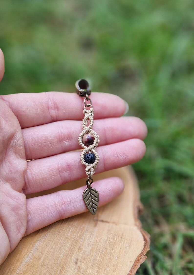 Puede incluir: Un pendiente de macram&eacute; hecho a mano con tres cuentas de piedra en tonos marr&oacute;n y negro. El pendiente presenta un dije de hoja y una parte superior de metal color bronce. El fondo est&aacute; borroso, con follaje verde y una superficie de madera.