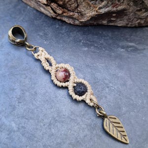 Puede incluir: Colgante de macram&eacute; hecho a mano con cierre y dije de hoja de lat&oacute;n. El colgante presenta un dise&ntilde;o tejido beige con una piedra rosa y marr&oacute;n y una piedra negra. El colgante mide aproximadamente 10 cm de largo.