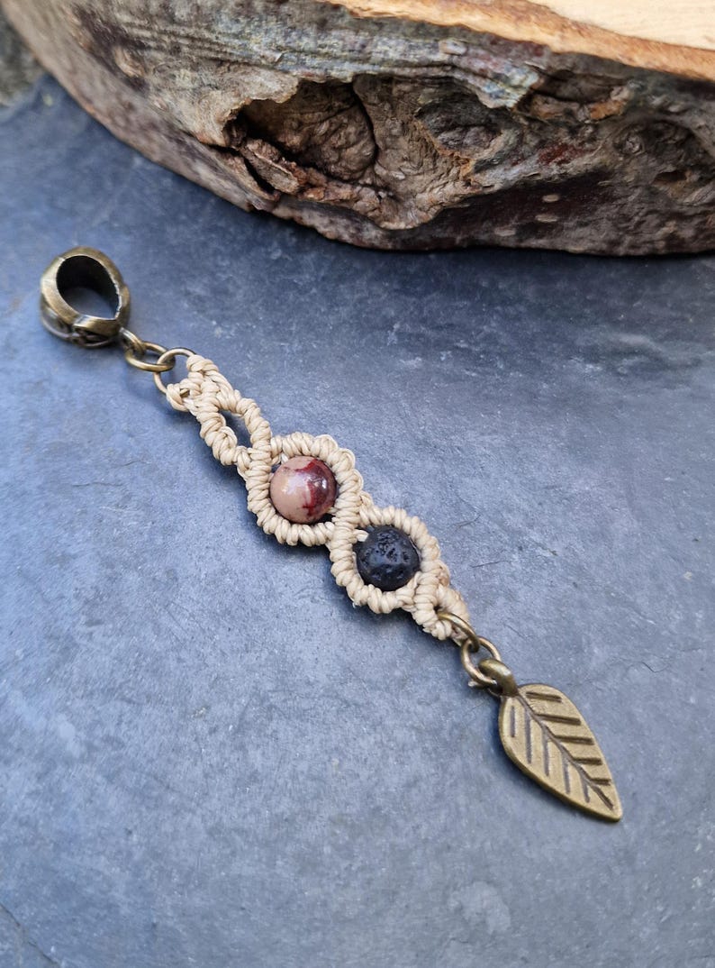 Puede incluir: Colgante de macram&eacute; hecho a mano con cierre y dije de hoja de lat&oacute;n. El colgante presenta un dise&ntilde;o tejido beige con una piedra rosa y marr&oacute;n y una piedra negra. El colgante mide aproximadamente 10 cm de largo.