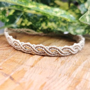 May include: A beige macrame bracelet with a woven design. The bracelet is made of a natural fiber and features a repeating pattern of connected ovals. The bracelet is displayed on a wooden surface with green foliage in the background.