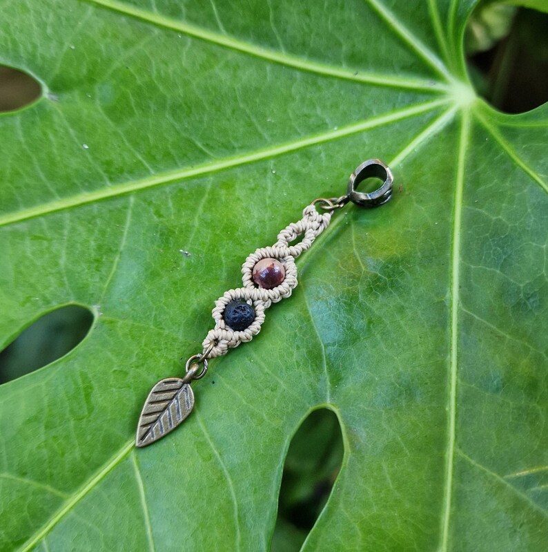 Puede incluir: Un colgante de macram&eacute; con tres cuentas de piedras preciosas en tonos marr&oacute;n, negro y rosa. Tiene un dije de hoja de bronce y un lazo para colgar. El colgante se muestra sobre una gran hoja verde.