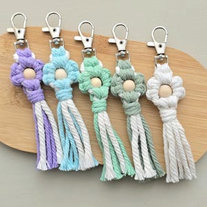 May include: Five macrame keyrings in pastel shades: purple, blue, green, and white. Each keyring has a flower design with a wooden bead centre and a tassel. The keyrings are displayed on a wooden surface.
