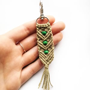 May include: A macrame keychain with a silver clasp, featuring a tan woven design with three green beads. The keychain has a tassel at the bottom, and is held in a hand.