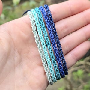 Puede incluir: Se muestran seis pulseras de macramé en tonos azules y verdes. Las pulseras están dispuestas en un degradado de verde claro a azul oscuro. Las pulseras están hechas de cordón tejido y son adecuadas para el uso diario.