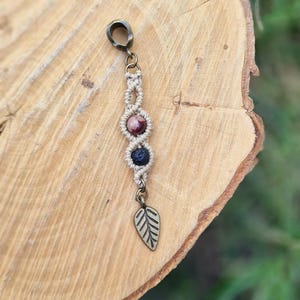 Puede incluir: Un charm de macram&eacute; hecho a mano con tres cuentas de piedras preciosas en tonos rosa, morado y azul. El charm est&aacute; acentuado con un charm de hoja de metal y un lazo de metal para colgar. El charm se muestra sobre una superficie de madera.