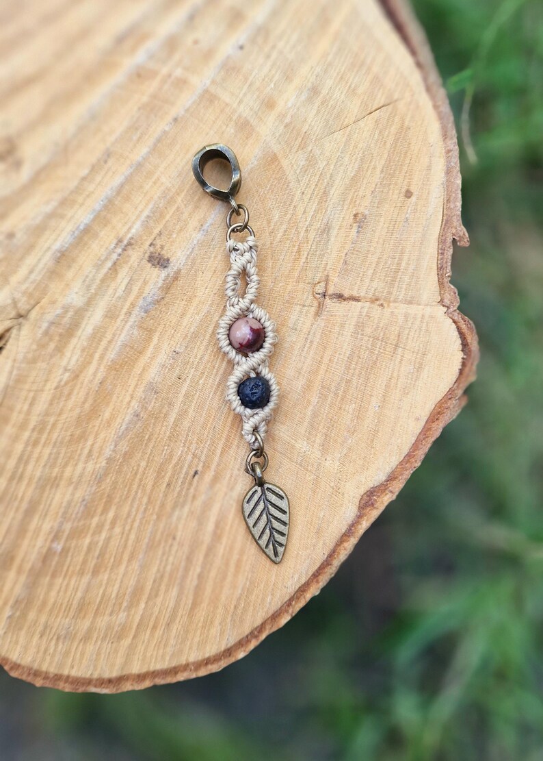 Puede incluir: Un charm de macram&eacute; hecho a mano con tres cuentas de piedras preciosas en tonos rosa, morado y azul. El charm est&aacute; acentuado con un charm de hoja de metal y un lazo de metal para colgar. El charm se muestra sobre una superficie de madera.