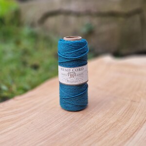 May include: A spool of teal hemp cord sits on a wooden surface. The label reads "HEMP CORD" with the brand name "Hemptique". The cord is tightly wound, showcasing its texture and color. The background is blurred, highlighting the cord.