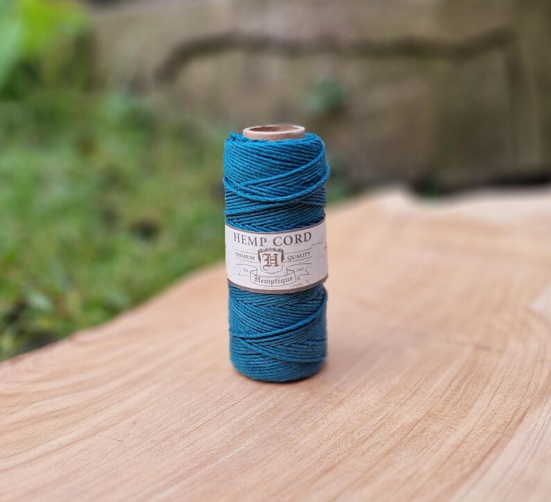 May include: A spool of teal hemp cord sits on a wooden surface. The label reads "HEMP CORD" with the brand name "Hemptique". The cord is tightly wound, showcasing its texture and color. The background is blurred, highlighting the cord.