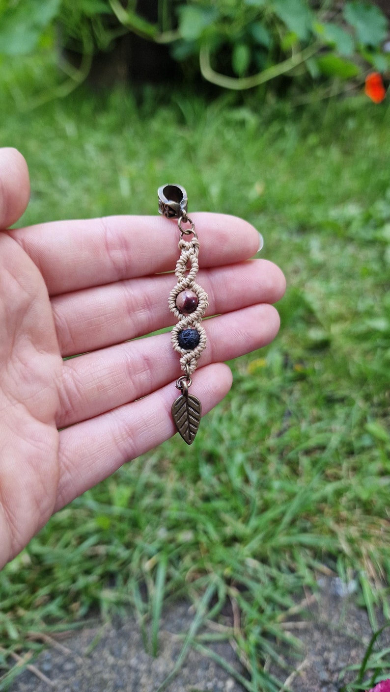 Puede incluir: Un charm de macram&eacute; hecho a mano con un cierre y un dije de hoja de color bronce. El charm presenta tres secciones decorativas con cuentas de piedra natural. Las cuentas son una mezcla de marr&oacute;n y negro, conectadas por nudos de macram&eacute; beige.
