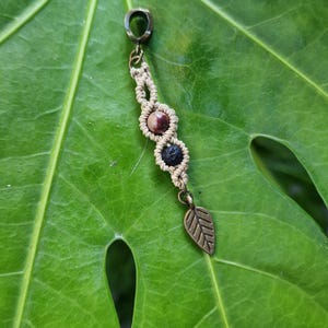 Puede incluir: Un colgante de macram&eacute; con un dije de hoja de bronce y tres cuentas: una piedra marr&oacute;n y blanca, y una cuenta de lava negra. El colgante est&aacute; suspendido de un anillo de bronce. El colgante est&aacute; sobre una hoja verde.