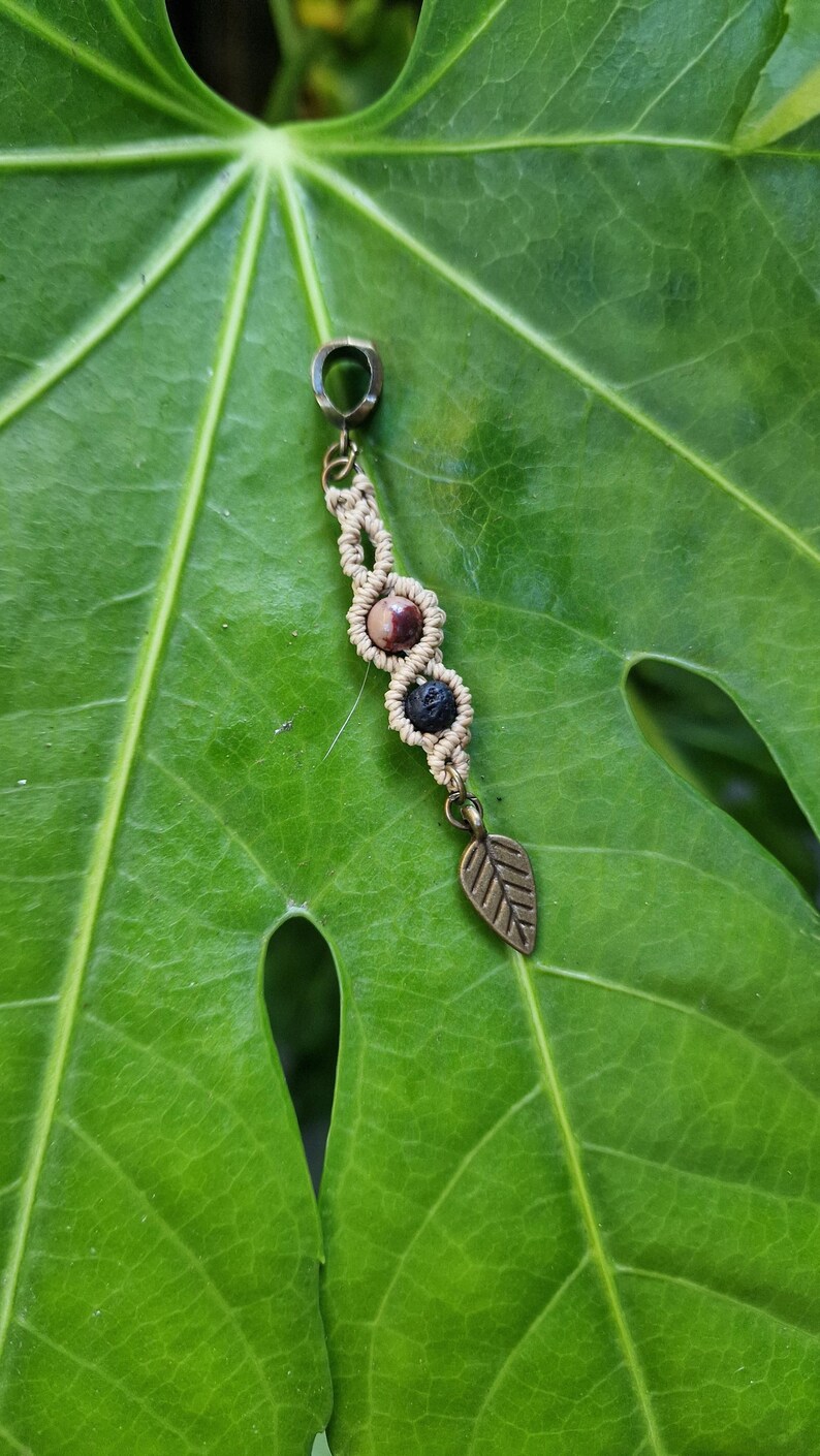 Puede incluir: Un colgante de macram&eacute; con un dije de hoja de bronce y tres cuentas: una piedra marr&oacute;n y blanca, y una cuenta de lava negra. El colgante est&aacute; suspendido de un anillo de bronce. El colgante est&aacute; sobre una hoja verde.