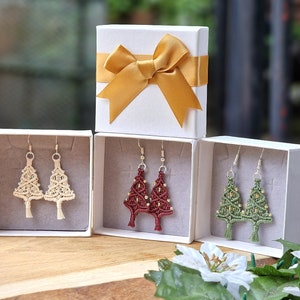May include: Earrings in white gift boxes. The earrings are shaped like Christmas trees, with three different color options: beige, burgundy, and green. A white gift box with a gold bow is also present, suggesting a holiday theme.