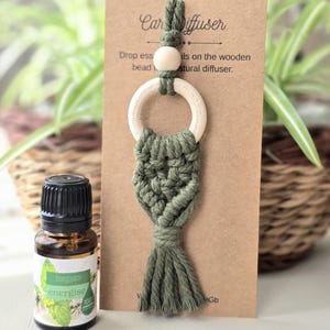 Może przedstawiać: Zielony dyfuzor samochodowy z makramy z drewnianym pierścieniem i koralikiem, zawieszony na brązowej karcie. Po lewej stronie mała brązowa szklana butelka olejku eterycznego "energise". Karta zawiera napis "Dyfuzor samochodowy" i instrukcje.