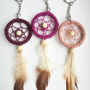 May include: Three dreamcatcher keychains in shades of burgundy, magenta, and peach. Each features a woven circular frame with a central bead, dangling feathers, and a silver keyring. The feathers transition from white to brown.