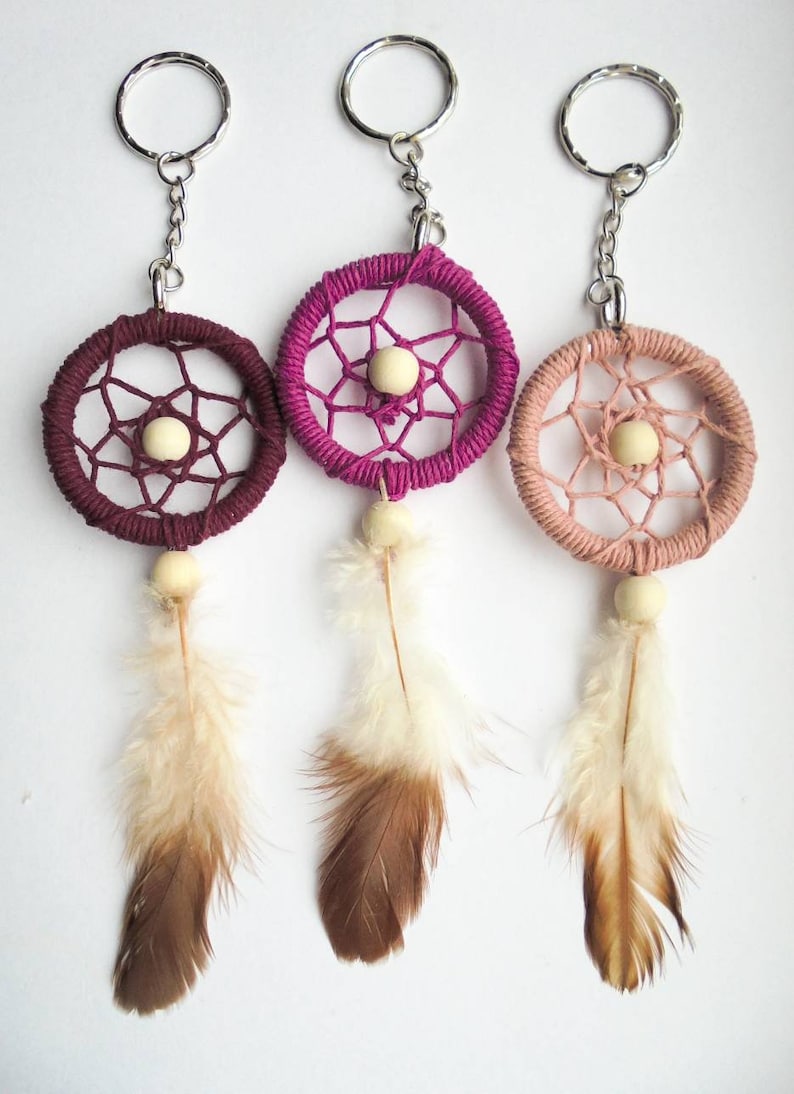 May include: Three dreamcatcher keychains in shades of burgundy, magenta, and peach. Each features a woven circular frame with a central bead, dangling feathers, and a silver keyring. The feathers transition from white to brown.