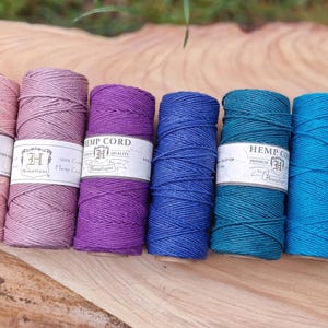 May include: A collection of colorful hemp cord spools in shades of pink, purple, blue, teal, and green. Each spool has a white label with the text "HEMP CORD". The spools are arranged on a wooden surface.