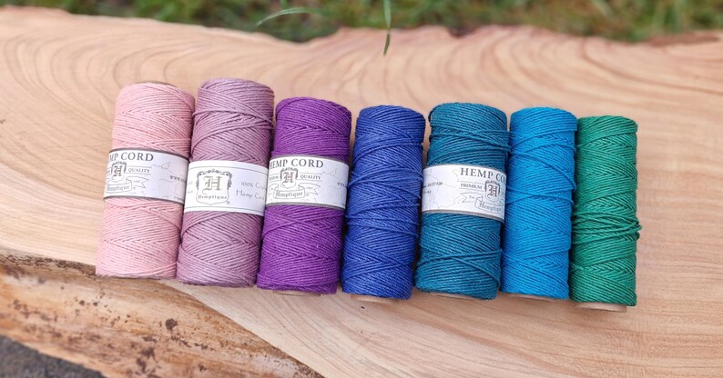 May include: A collection of colorful hemp cord spools in shades of pink, purple, blue, teal, and green. Each spool has a white label with the text "HEMP CORD". The spools are arranged on a wooden surface.