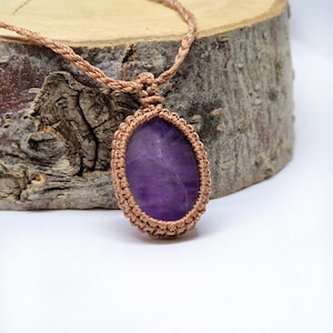 Puede incluir: Un collar de macramé artesanal con una gran amatista ovalada. La piedra púrpura está engastada en un marco de macramé tejido de color marrón claro y suspendida de un cordón trenzado a juego. El collar se muestra sobre un fondo de madera natural.