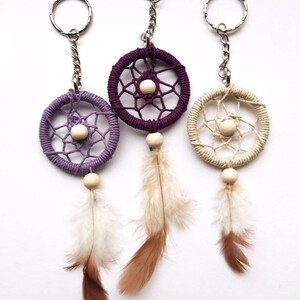 May include: Three dreamcatcher keychains in purple, beige, and cream colors. Each keychain features a woven circle with a wooden bead center, hanging from a silver chain and ring. Feathers in shades of brown and white dangle below.