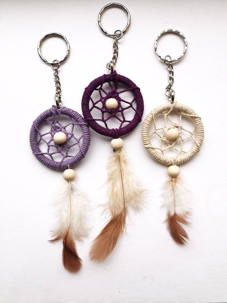 May include: Three dreamcatcher keychains in purple, beige, and cream colors. Each keychain features a woven circle with a wooden bead center, hanging from a silver chain and ring. Feathers in shades of brown and white dangle below.