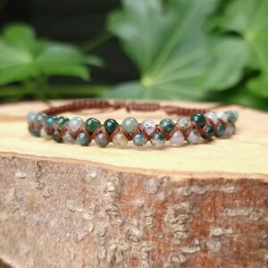Puede incluir: Una pulsera de doble hebra con cuentas moteadas verdes y blancas, ensartadas en un cordón marrón. Las cuentas están dispuestas en un patrón repetitivo, creando un aspecto texturizado. La pulsera se exhibe sobre una superficie de madera, con follaje verde en el fondo.