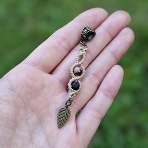 Puede incluir: Un pendiente de macram&eacute; hecho a mano con un dije de hoja de bronce. El pendiente presenta tres cuentas en tonos marr&oacute;n y negro, conectadas por un cord&oacute;n beige tejido. La parte superior tiene una tapa decorativa de bronce.