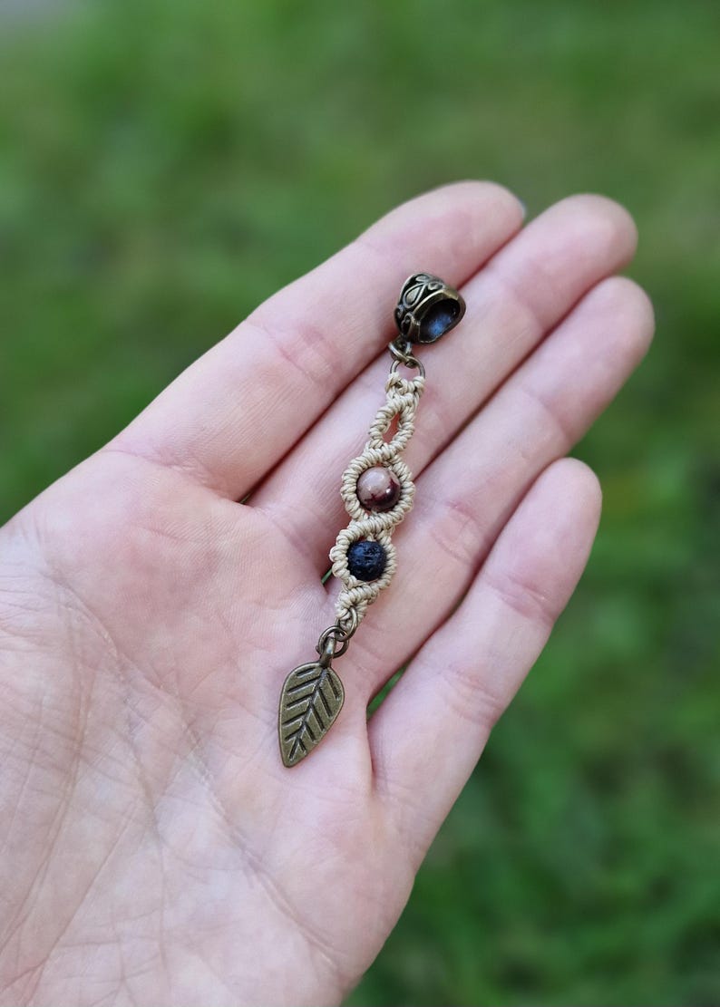 Puede incluir: Un pendiente de macram&eacute; hecho a mano con un dije de hoja de bronce. El pendiente presenta tres cuentas en tonos marr&oacute;n y negro, conectadas por un cord&oacute;n beige tejido. La parte superior tiene una tapa decorativa de bronce.