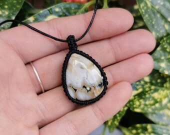 May include: A teardrop-shaped pendant necklace featuring a cream-colored stone with brown and black veining. The stone is encased in a black macrame setting and hangs from a black cord. The necklace is displayed against a backdrop of green foliage.