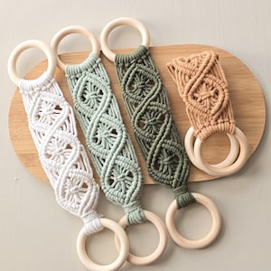 May include: Four macrame straps featuring intricate knotted patterns in white, sage green, olive green, and tan. Each strap is attached to two smooth, circular wooden rings at both ends, creating a functional design with a textured cord finish.