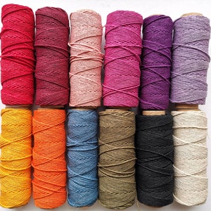 May include: A collection of colorful twine spools in various shades, including red, pink, purple, yellow, orange, blue, black, and beige. The twine is tightly wound on cardboard spools, ideal for crafting and decorative projects.