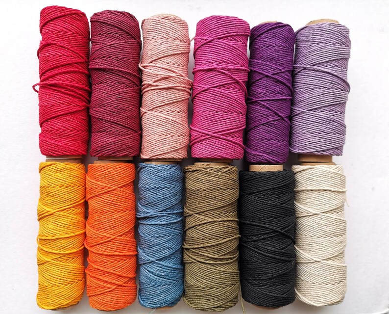 May include: A collection of colorful twine spools in various shades, including red, pink, purple, yellow, orange, blue, black, and beige. The twine is tightly wound on cardboard spools, ideal for crafting and decorative projects.