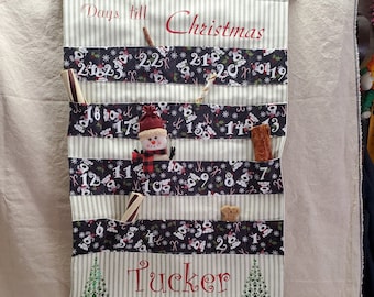 May include: A white and green striped fabric advent calendar with the text "Days till Christmas" and "Tucker" in red. The calendar has pockets with numbers 1-23 and contains dog treats and toys. A snowman, a green gift, and a gingerbread man are in front of the calendar.