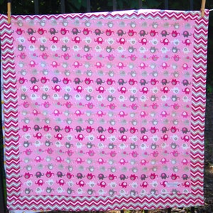 May include: A pink baby blanket with a repeating pattern of colorful elephants. The blanket has a pink and white chevron border. The elephants are in shades of pink, gray, and white.