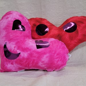May include: Two heart-shaped pillows, one pink with a crescent moon and two hearts, the other red with a circle and a heart, all with a shiny purple finish.