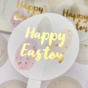May include: Round stickers with gold foil text that reads "Happy Easter." The stickers feature watercolor-style Easter eggs in pastel colors. The eggs are pink, beige, and green with speckled details. The stickers are on a white background.