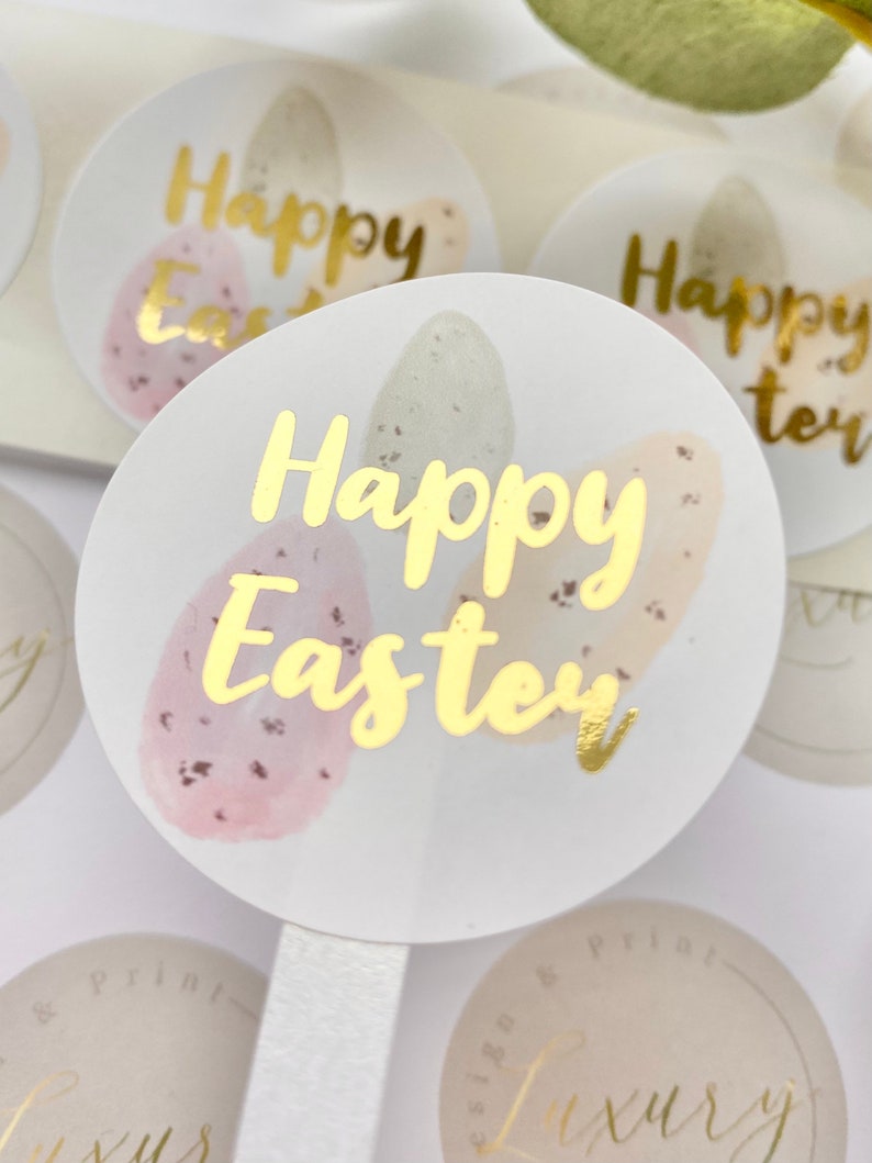 May include: Round stickers with gold foil text that reads "Happy Easter." The stickers feature watercolor-style Easter eggs in pastel colors. The eggs are pink, beige, and green with speckled details. The stickers are on a white background.
