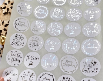 May include: A sheet of silver and white Christmas-themed stickers. Each sticker is round and features festive phrases like "Merry Christmas," "Let it Snow," and "Santa Baby." Some stickers have snowflake, snowman, or reindeer designs.