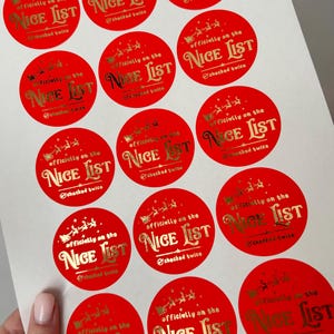 May include: A sheet of red, circular stickers with gold foil text. Each sticker says "Officially on the Nice List, checked twice" with a Santa and sleigh graphic. Suitable for festive gift wrapping or crafting.