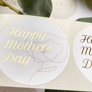 Puede incluir: Primer plano de pegatinas redondas con letras doradas. Las pegatinas dicen "Happy Mothers Day" y presentan un delicado contorno de rosa. El fondo es de un color suave y neutro, con toques de vegetaci&oacute;n.