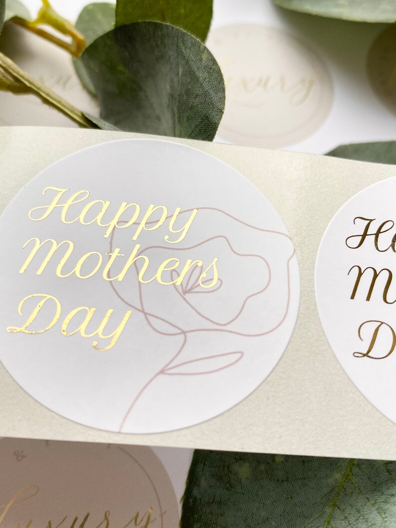 Puede incluir: Primer plano de pegatinas redondas con letras doradas. Las pegatinas dicen "Happy Mothers Day" y presentan un delicado contorno de rosa. El fondo es de un color suave y neutro, con toques de vegetaci&oacute;n.