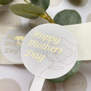 Puede incluir: Pegatinas redondas con texto dorado "Happy Mothers Day" y un dise&ntilde;o floral. Las pegatinas son de color gris claro con una cinta blanca y est&aacute;n rodeadas de hojas verdes.