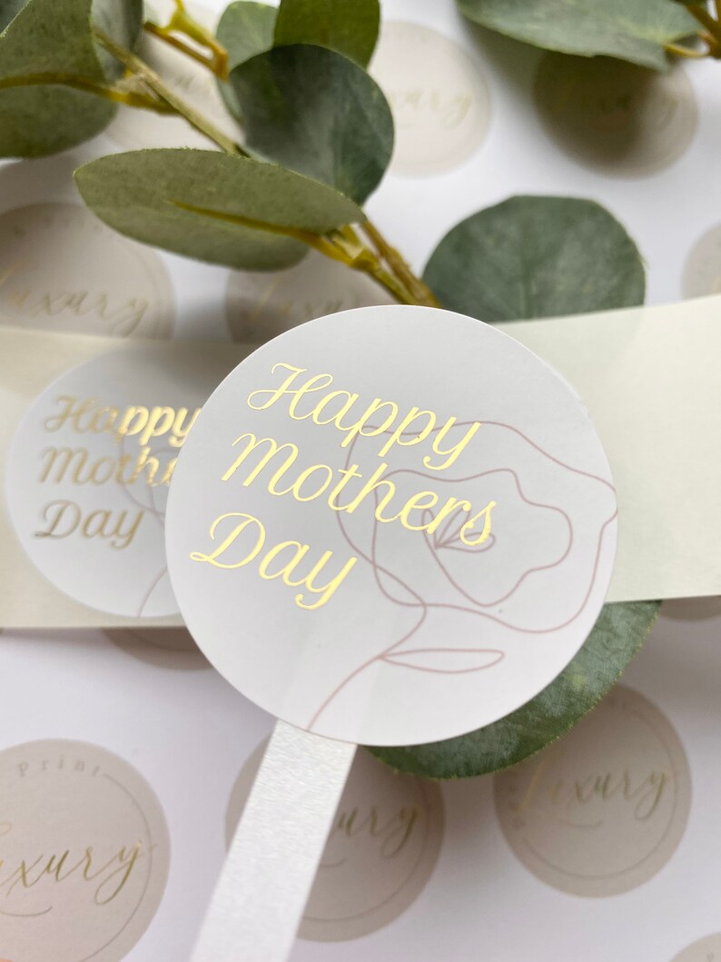 Puede incluir: Pegatinas redondas con texto dorado "Happy Mothers Day" y un dise&ntilde;o floral. Las pegatinas son de color gris claro con una cinta blanca y est&aacute;n rodeadas de hojas verdes.