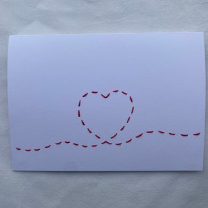 May include: A white card with a red stitched heart design. The heart is outlined with a dashed line and the stitching continues in a wavy line across the bottom of the card.