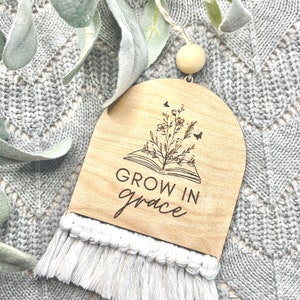 May include: Wooden sign with a white tassel, featuring a book with flowers and butterflies, and the text "GROW IN grace".